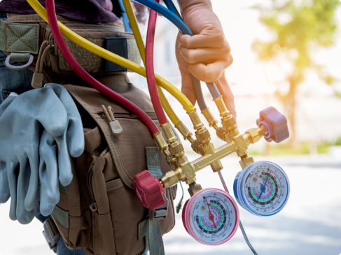 Person holding HVAC manifold gauge set with colored hoses, next to a tool pouch with gloves attached, outdoors in daylight.